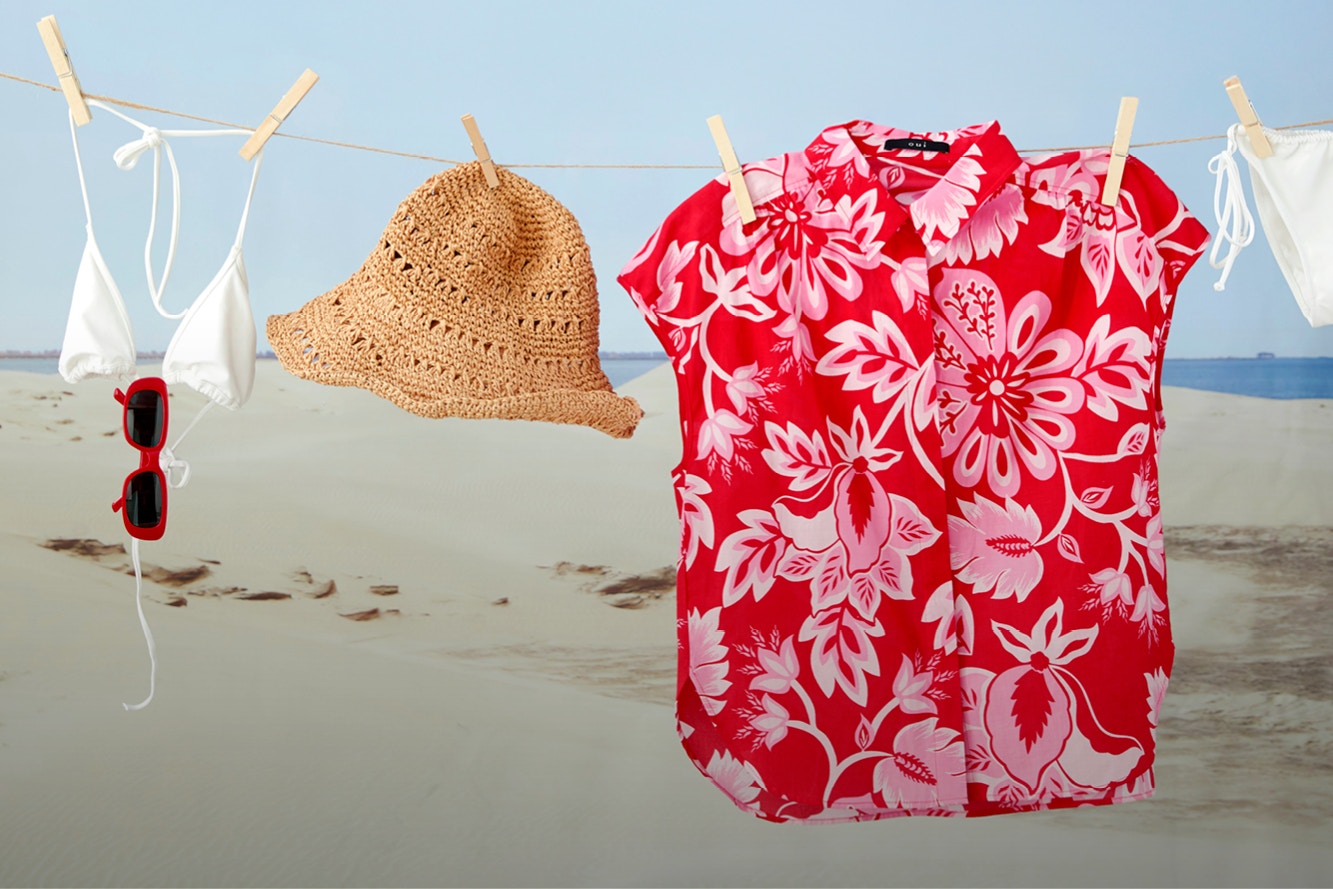 Eine Bluse in rot mit weissen Blumendruck, ein Bikinioberteil und Unterteil in weiss, ein Strohhut hängen auf einer Wäscheleine vor einer Strandkulisse.