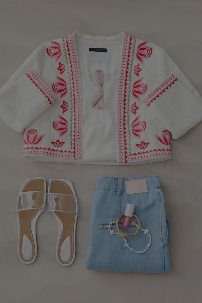 Ein Bild mit gelegter Bekleidung von oben Fotografiert. Eine weisse Jacke mit roten Ornamenten am Saum liegt oben und darunter links ein paar Sandalen und links eine hellblaue Hose.