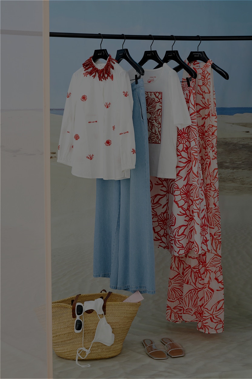 A white blouse with red shell elements, a denim skirt, a white shirt with a red front print and a skirt and maxi dress in white with a shell print hang on a pole in front of a beach backdrop.