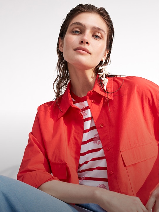 Woman wearing a red open blouse with a white and red striped shirt underneath and light blue jeans.