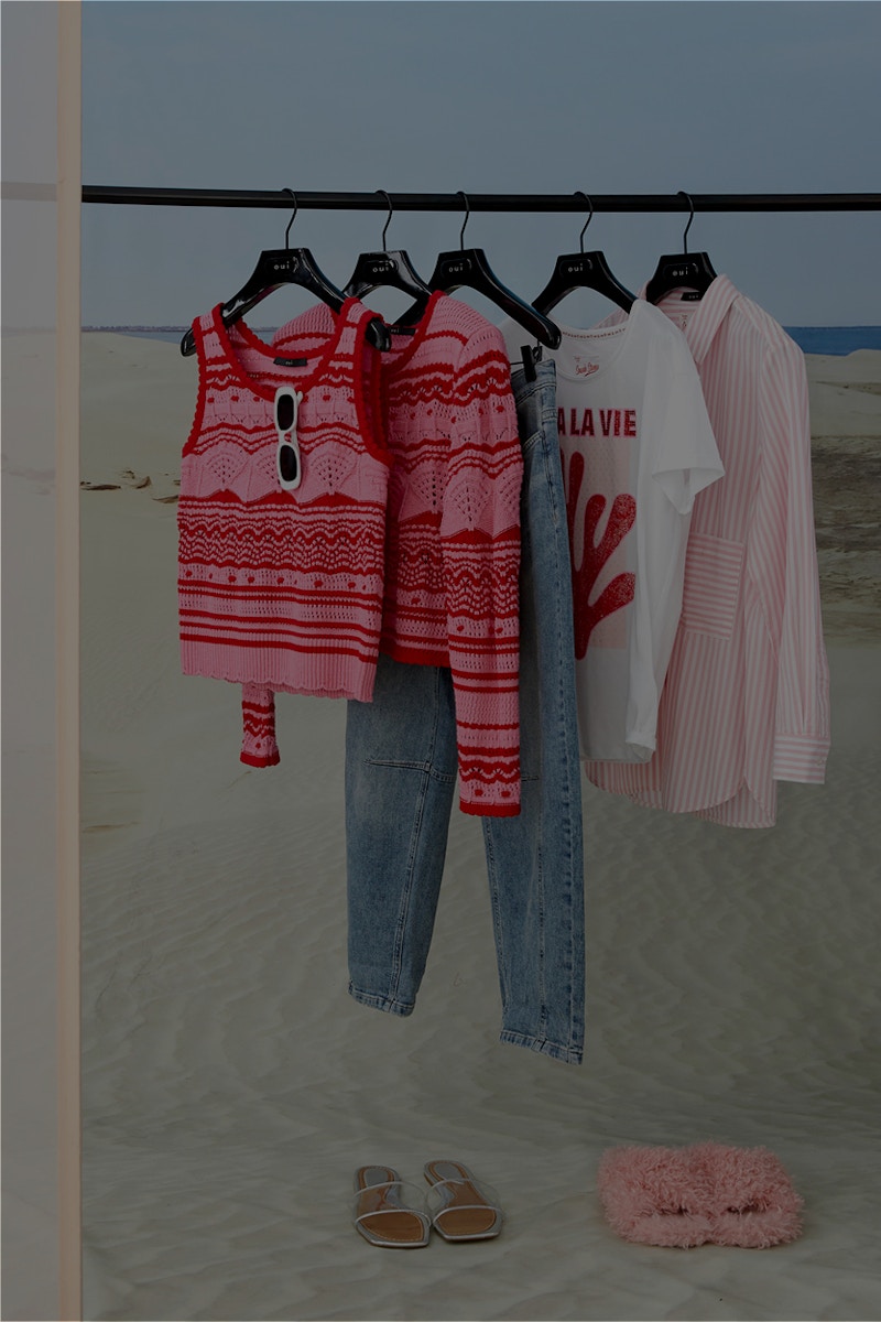 A pink and red striped knitted top and cardigan set, a pair of jeans, a white coral print shirt and a striped blouse hang on a pole in front of a beach backdrop.