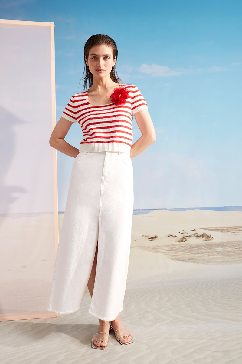 Woman wearing a red and white striped short-sleeved jumper with a red corsage and a white long, slit denim skirt.