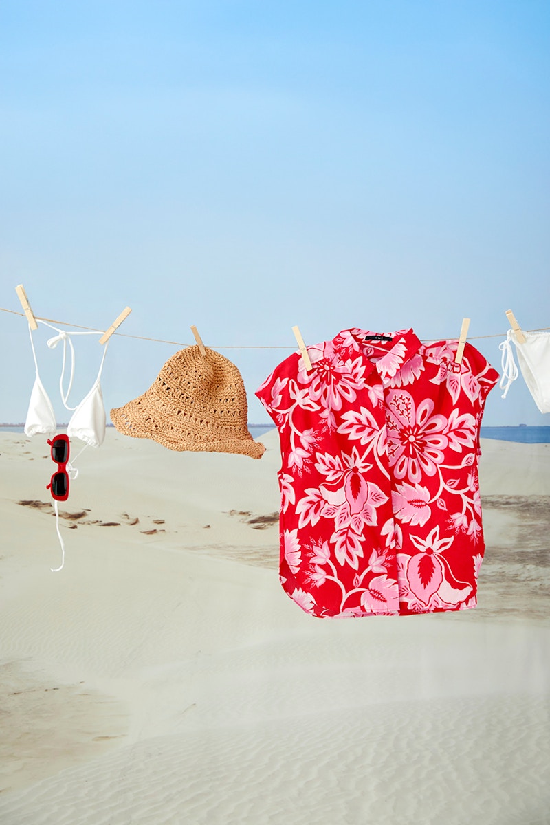 A blouse in red with a white floral print, a bikini top and bottom in white and a straw hat hanging on a washing line in front of a beach backdrop.
