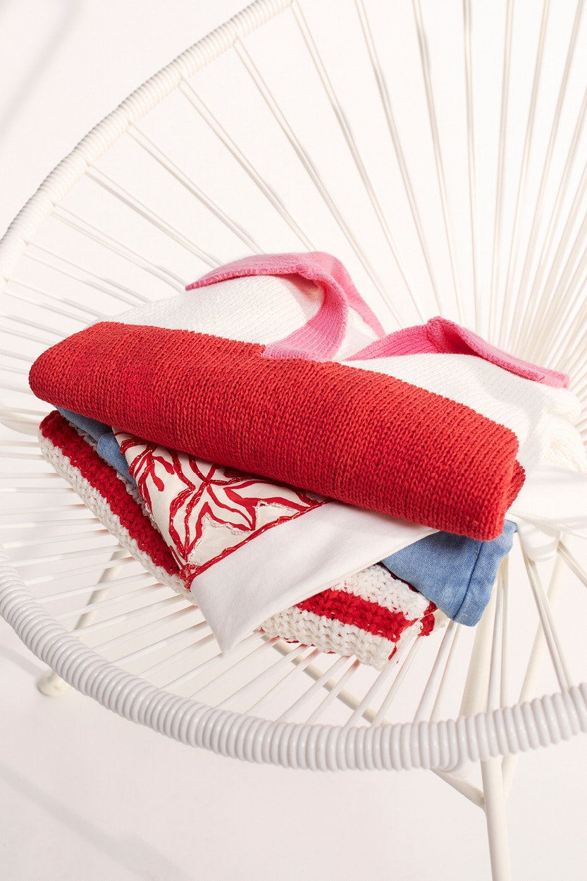 A pile of clothes lies on a white chair. You can see a red and white striped jumper with a pink polo collar, a white shirt with a red coral print, a pair of jeans and a red and white striped knitted jumper.