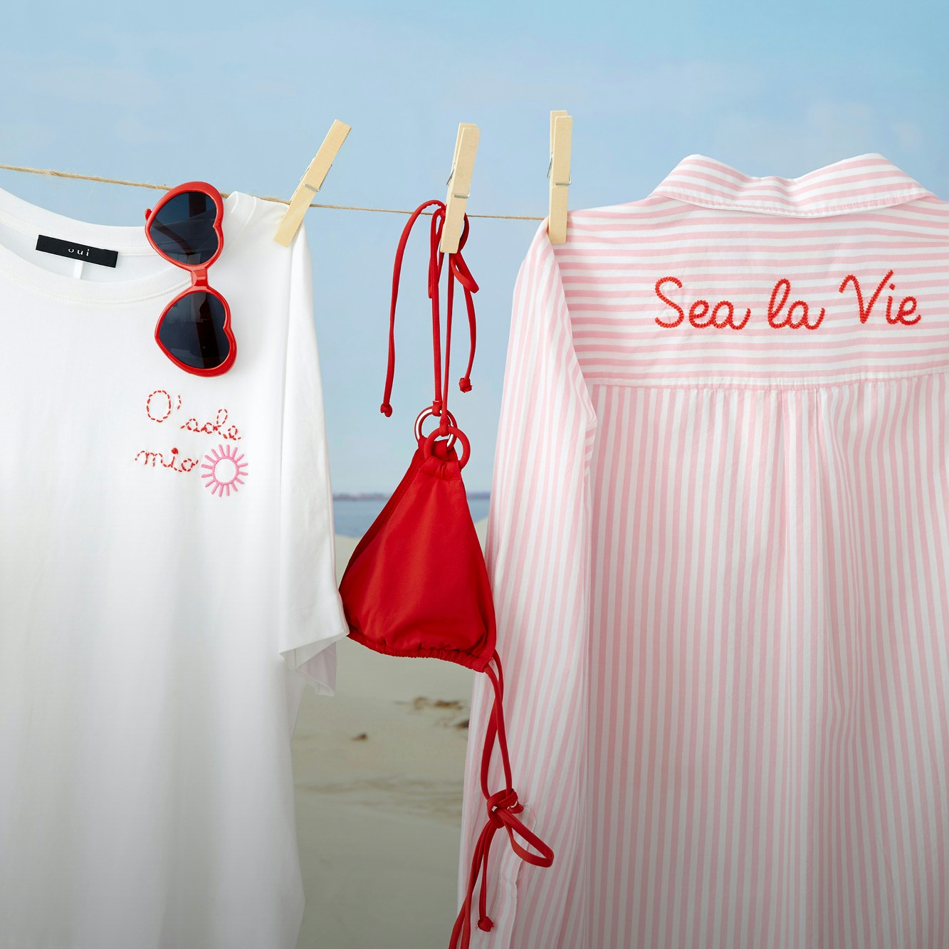 Eine Bluse in rot-weiss gestreift, ein Bikinioberteil in rot und ein T-shirt weiss mit rotem Lettering Druck hängen auf einer Wäscheleine vor einer Strandkulisse.