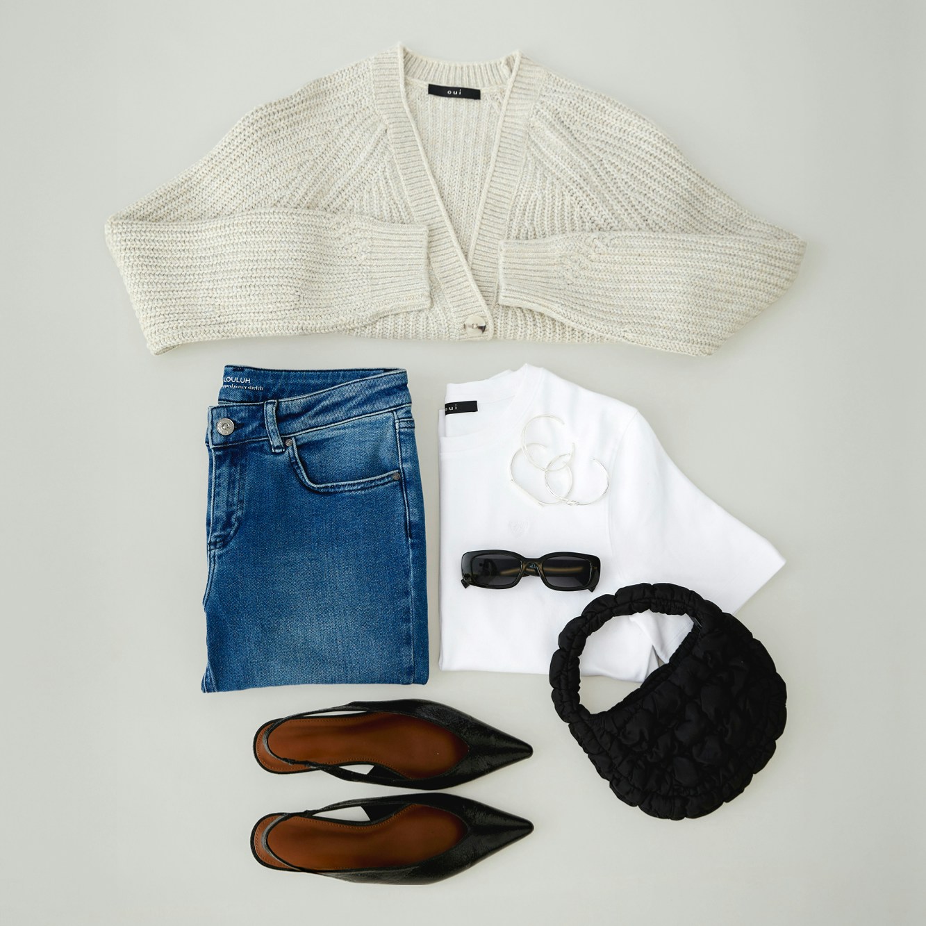 Ein Bild von oben auf gefaltete Kleidung. Oben ein beiger Cardigan darunter links eine gefaltete blaue Jeans und daneben rechts ein gefaltetes weißes T-Shirt, vervollständig wird es durch eine Sonnenbrille, Tasche und Sandalen.