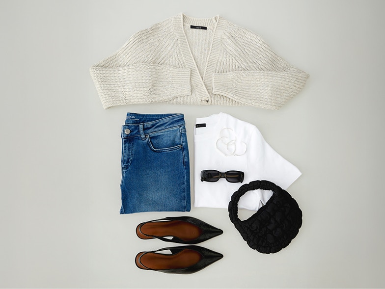 A picture from above on folded clothes. A beige cardigan on top, a folded pair of blue jeans on the left and a folded white T-shirt on the right, completed with sunglasses, a bag and sandals.