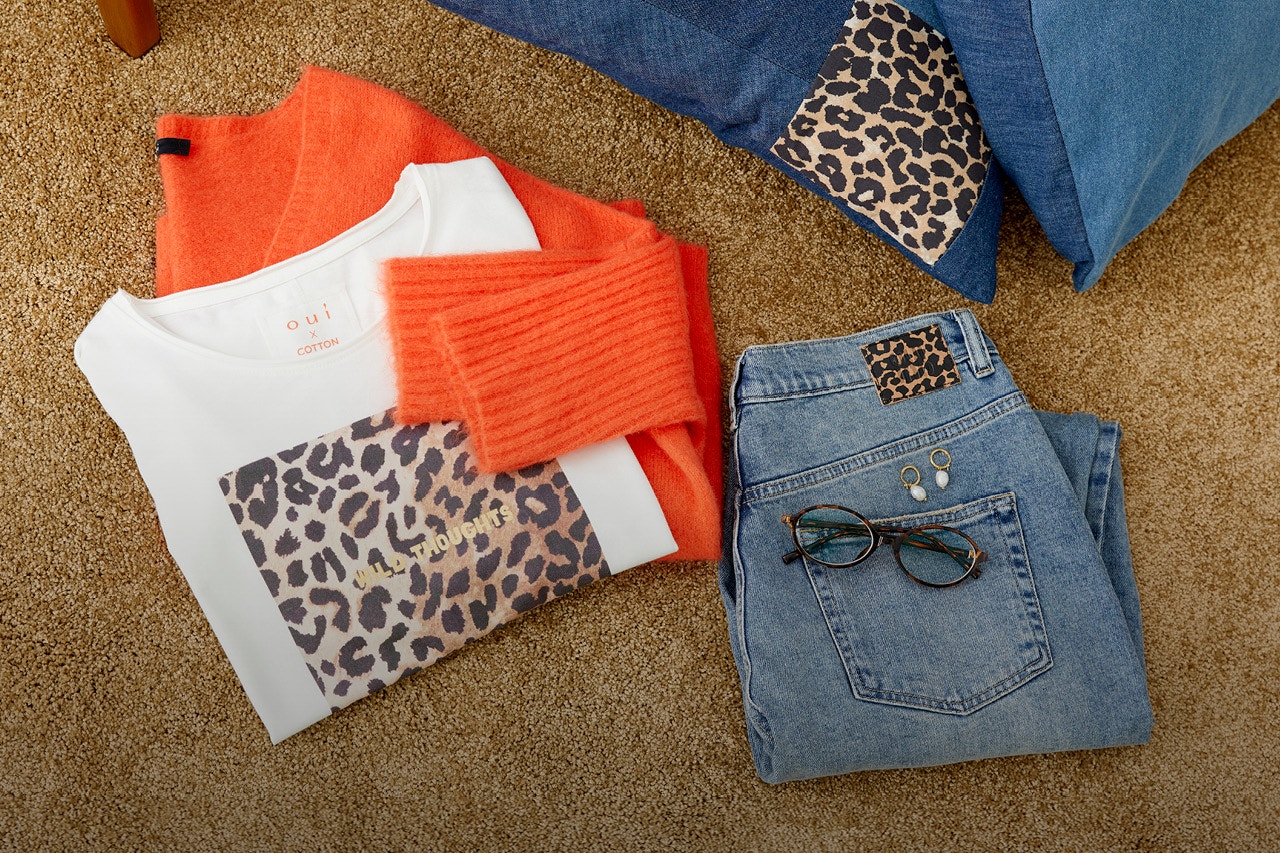 A picture with clothes laid out on a brown-beige carpet. You can see a folded T-shirt with a placed leopard print, behind it an orange jumper and some jeans.