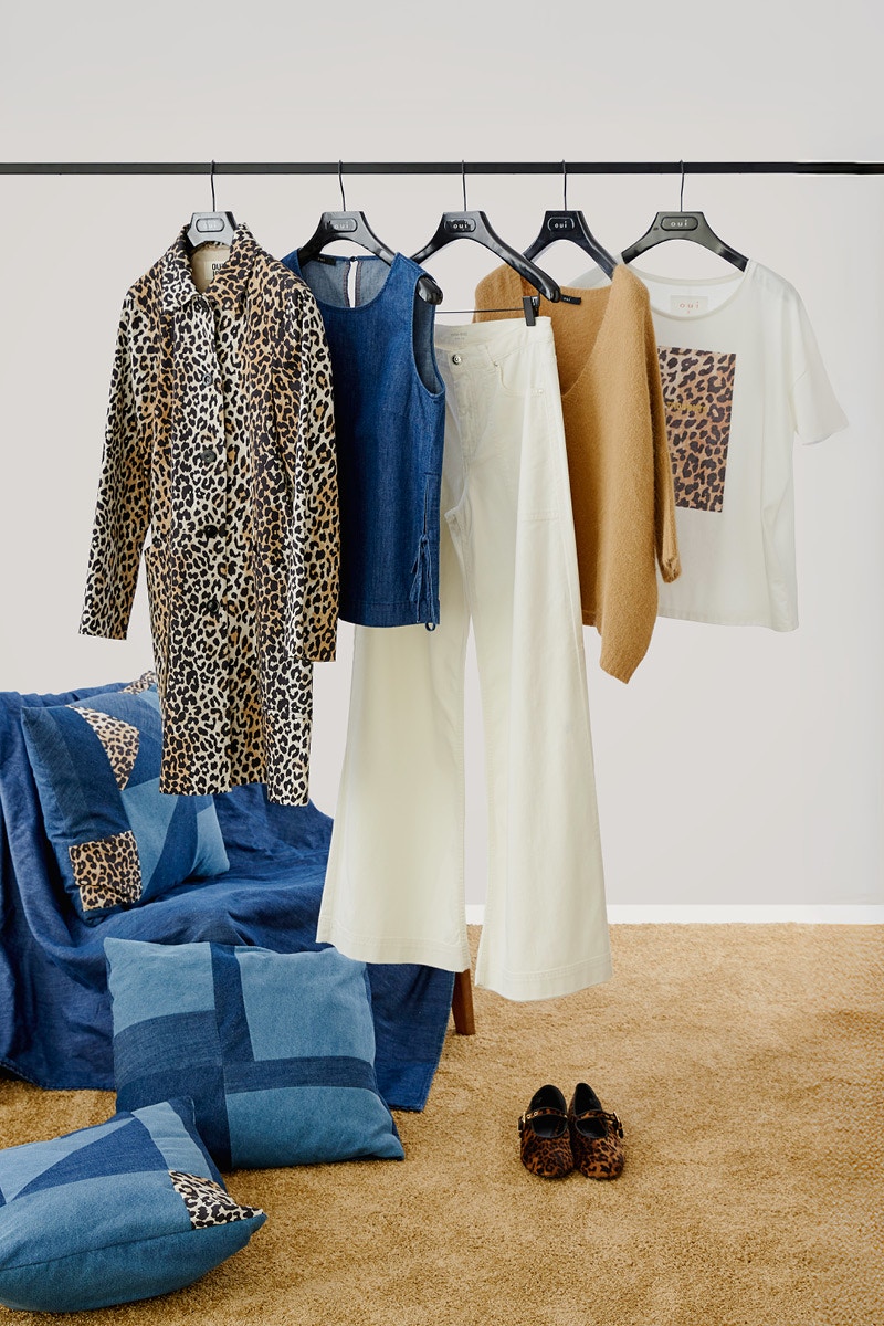 Clothes rail with hanging clothes next to a pile of denim and leo patch cushions. A leotard, denim top, white trousers, brown jumper and a white shirt with a leopard print on the front are hanging on the rail.