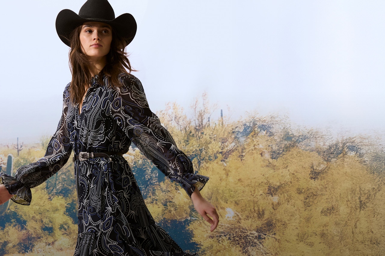 Woman with a black cowboy hat and a patterned dress in black with a belt.