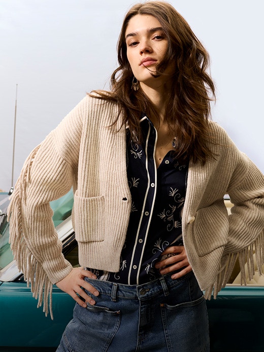 A woman is wearing a beige cardigan with fringes on the sleeves, a black blouse with a white print and jeans underneath. The look is reminiscent of a cowgirl.