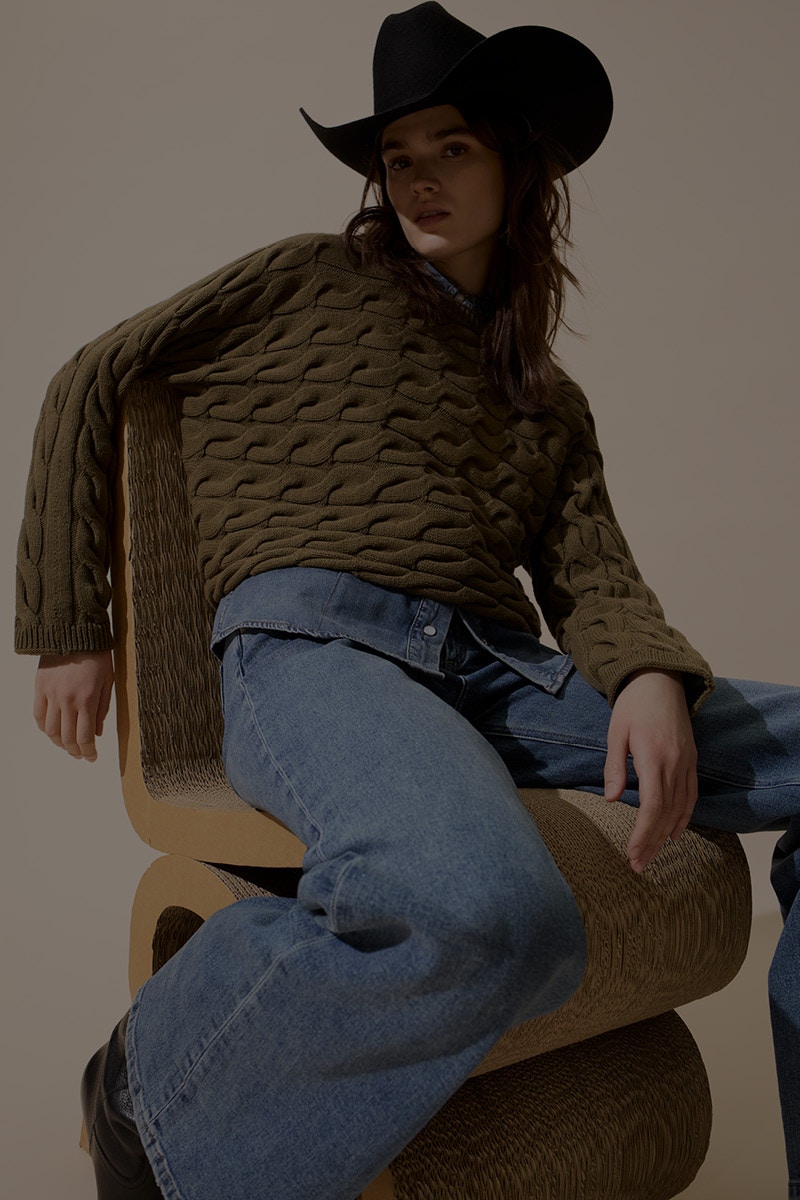 Woman on a chair wearing a black cowboy hat, a green knitted jumper, a denim shirt with a visible hem underneath and denim trousers.