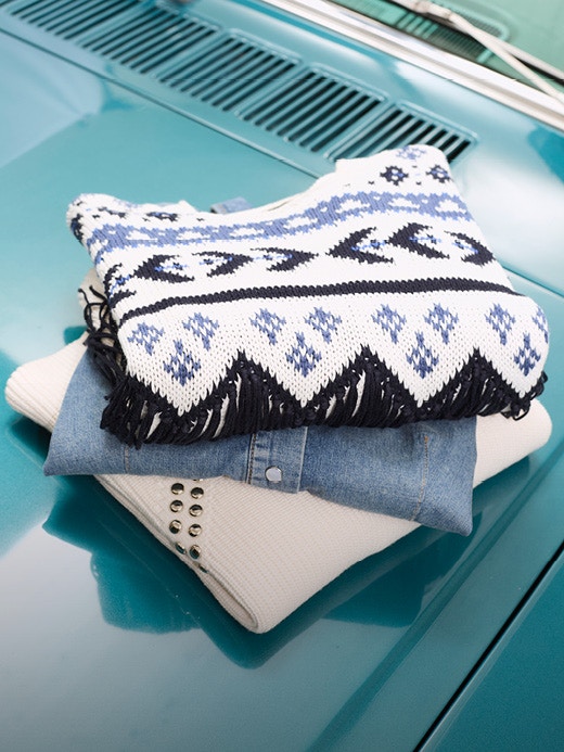 Ein Stapel gefalteter Kleidung auf einer türkisen Motorhaube. Ein Pullover weiss mit einem Ikat Muster in blau und schwarz, darunter ein Jeanshemd und da drunter ein beiger Pullover mit Nieten.