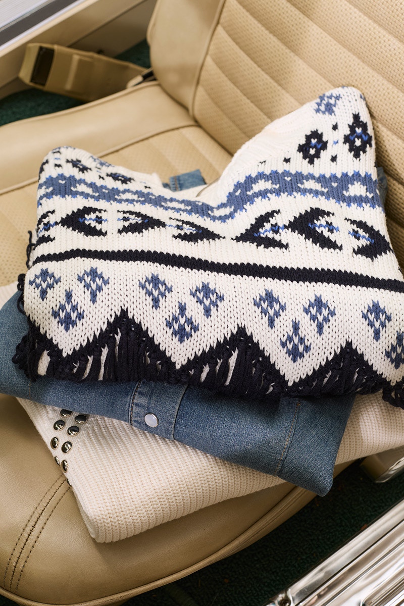 Ein Stapel gefalteter Kleidung auf einem Autositz. Ein Pullover weiss mit einem Ikat Muster in blau und schwarz, darunter ein Jeanshemd und da drunter ein beiger Pullover mit Nieten.