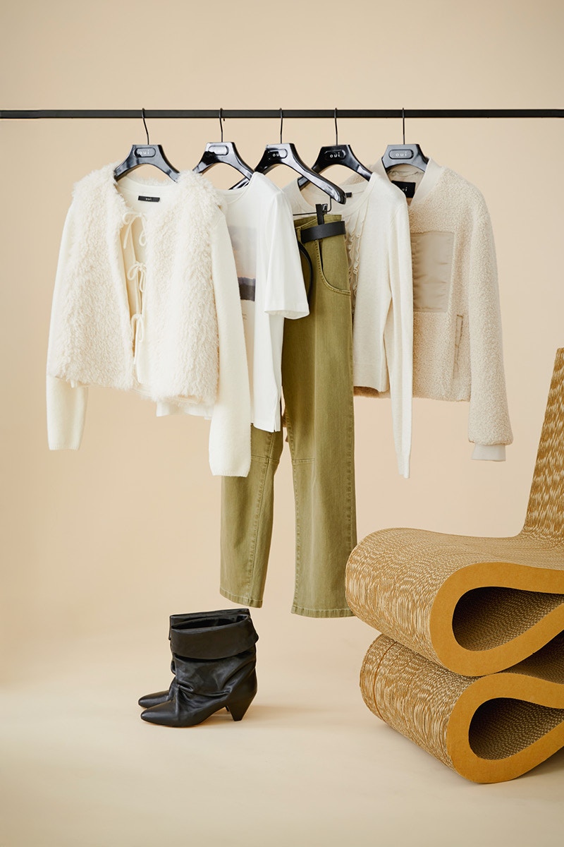 Kleidung hängt auf einer Stange. Erst eine weisse Weste mit einer Strickjacke in weiss darunter, dann ein weißes T-Shirt mit einem Aufdruck, eine gruene Hose, ein weißes Oberteil und eine beige Strickjacke.