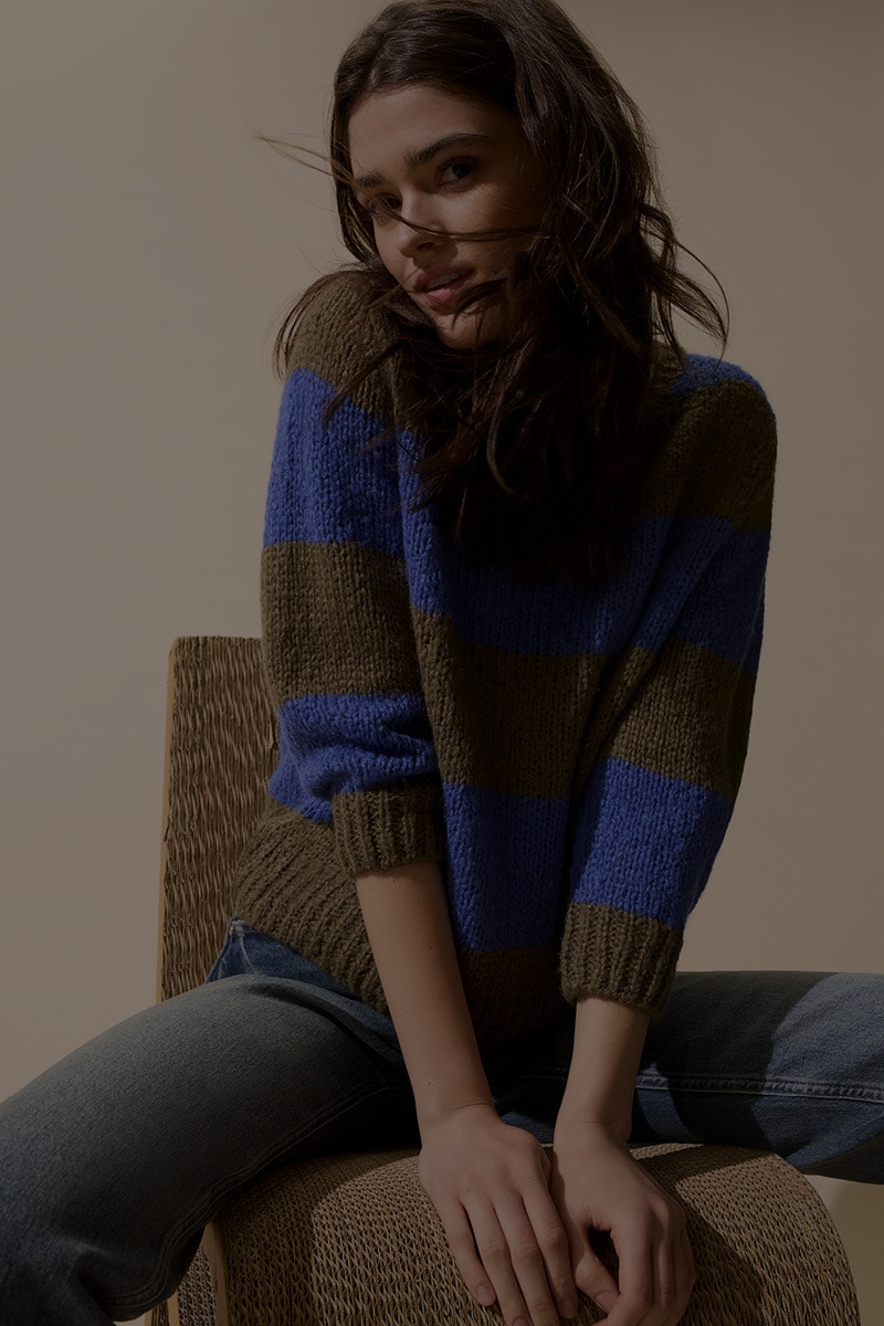 Frau auf einem Stuhl trägt einen gestreiften Blockstreifen Pullover in gruen mit blau und dazu eine Jeanshose.