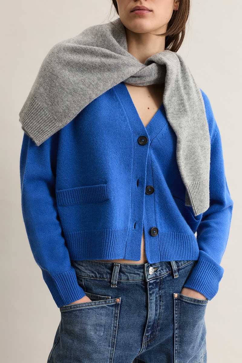 Frau mit einem blauen Cardigan und einer Jeans. Um die Schultern hat sie einen grauen Pullover oder Strickjacke gehängt.