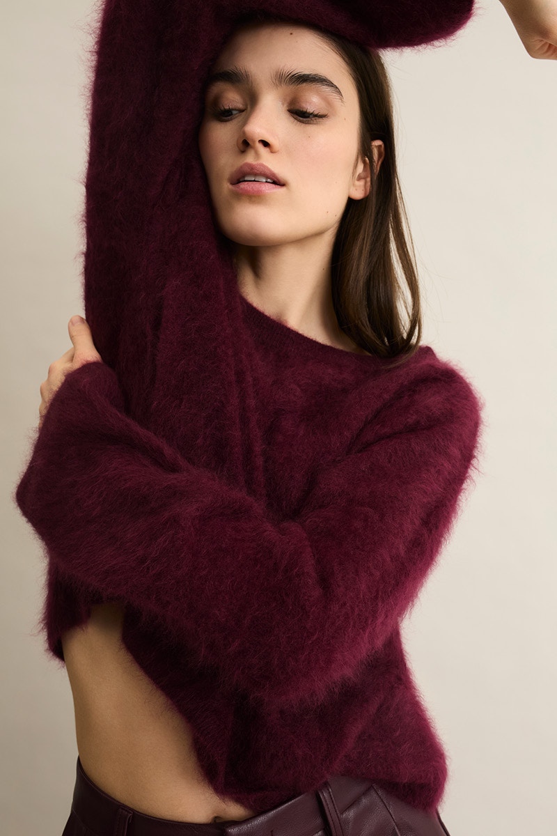 A woman with a burgundy red jumper. The left arm is placed over the head so that some belly skin is shown.