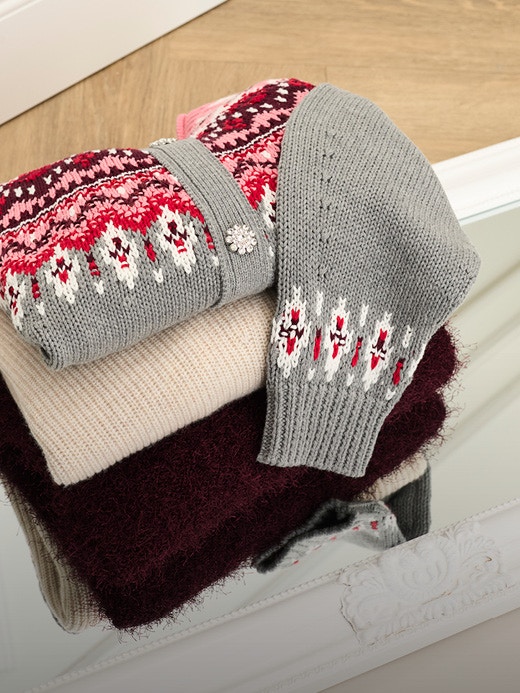 Ein Stapel gefalteter Strickteile, oben auf eine graue Strickjacke mit einem Norweger-Muster aus pink, rot und weiss, darunter ein beiger Pullover, darunter ein roter flauschiger Pullover.