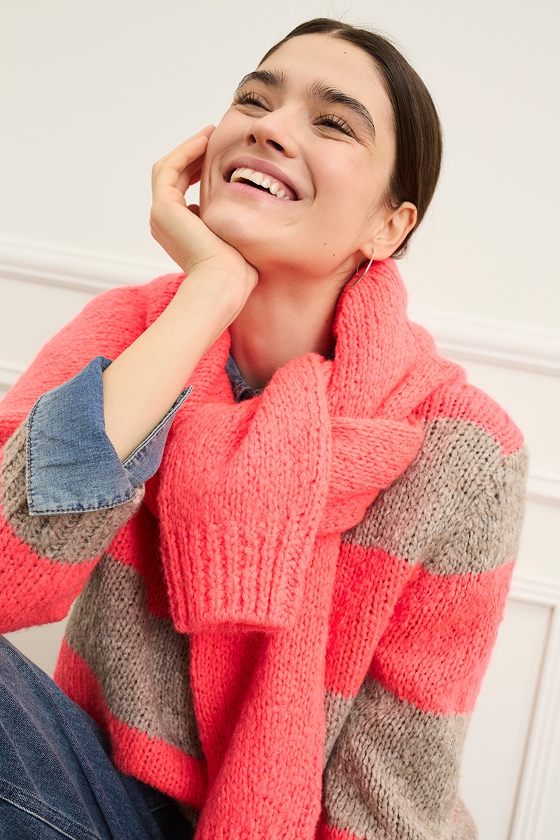 Lächelnde Frau trägt einen grau, pink gestreiften Pullover mit einem pinken Pullover über die Schultern und einer Jeans.