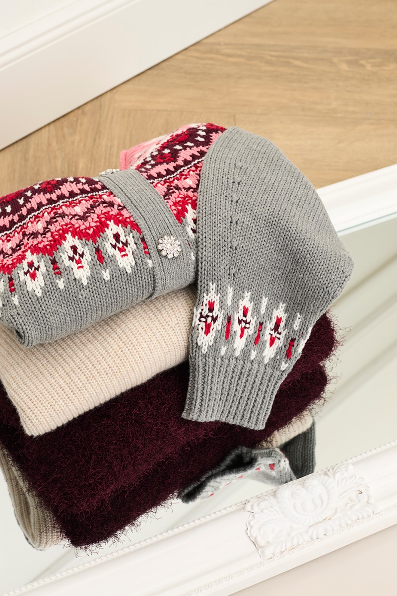 Ein Stapel gefalteter Strickteile, oben auf eine graue Strickjacke mit einem Norweger-Muster aus pink, rot und weiss, darunter ein beiger Pullover, darunter ein roter flauschiger Pullover.