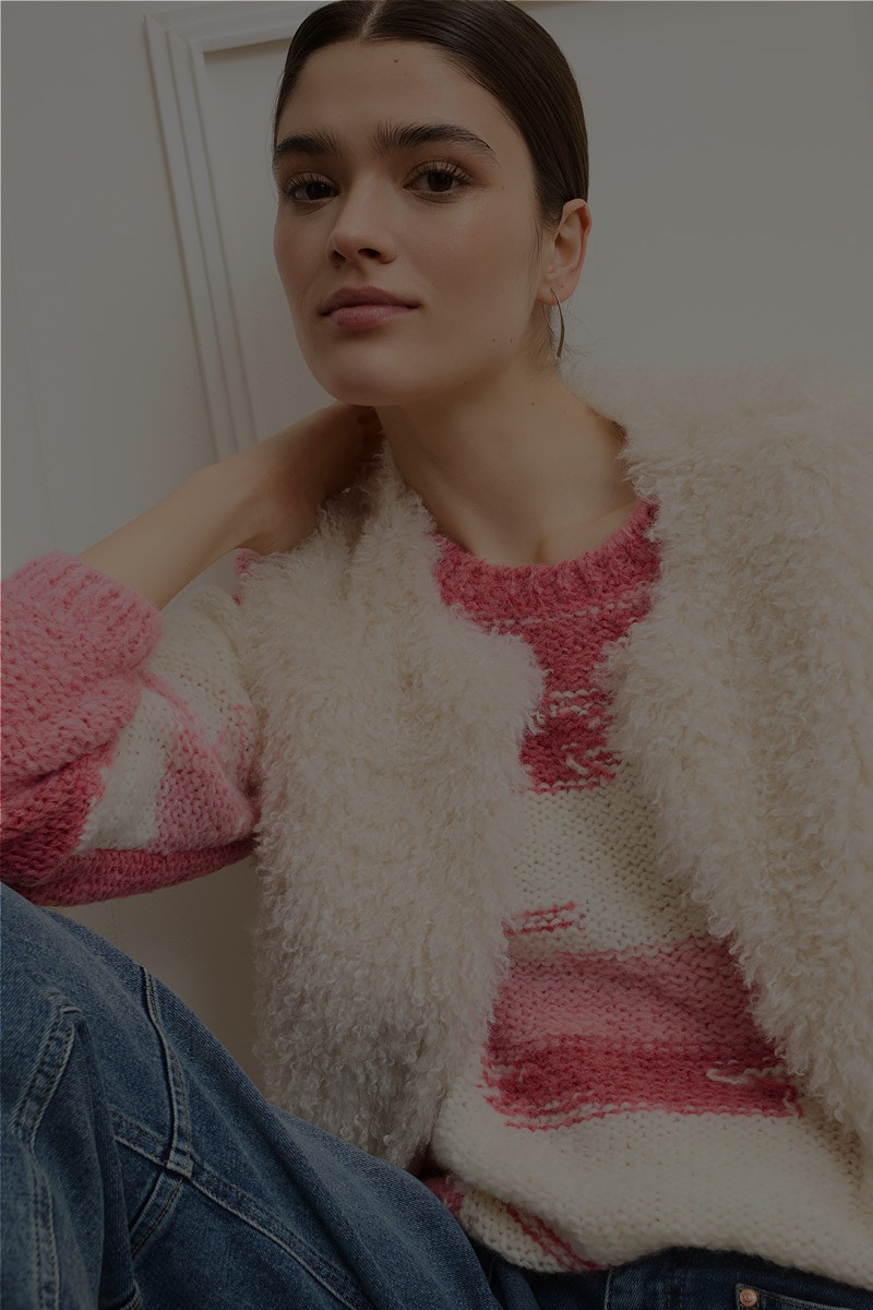 Frau mit einem weissen, pink, roten Pullover mit einer flauschigen weissen Weste darüber und dazu eine Jeans.