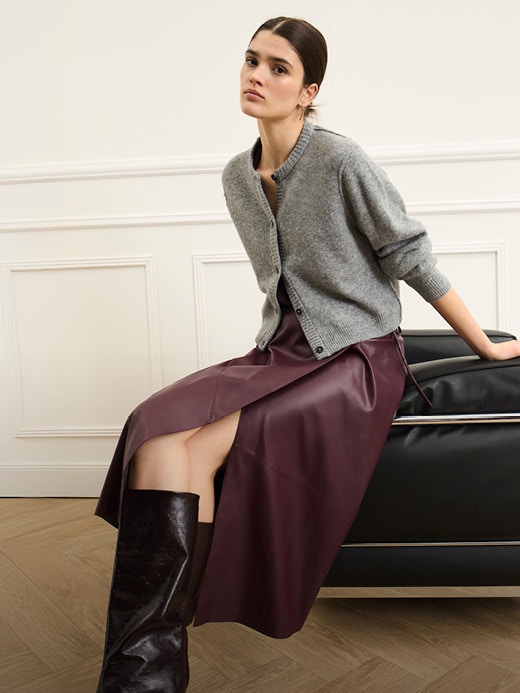 A woman sits at the corner of a black armchair. She is wearing a grey cardigan with a brown leather-look wrap skirt.