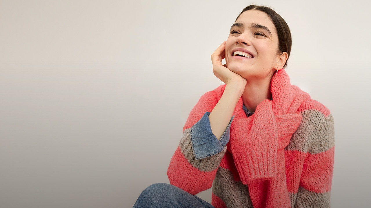 Lächelnde Frau trägt einen grau, pink gestreiften Pullover mit einem pinken Pullover über die Schultern.