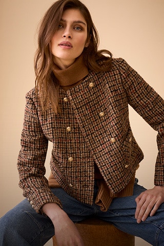 Woman sitting wearing a brown checked jacket with gold buttons, underneath a brown turtleneck jumper and jeans trousers.