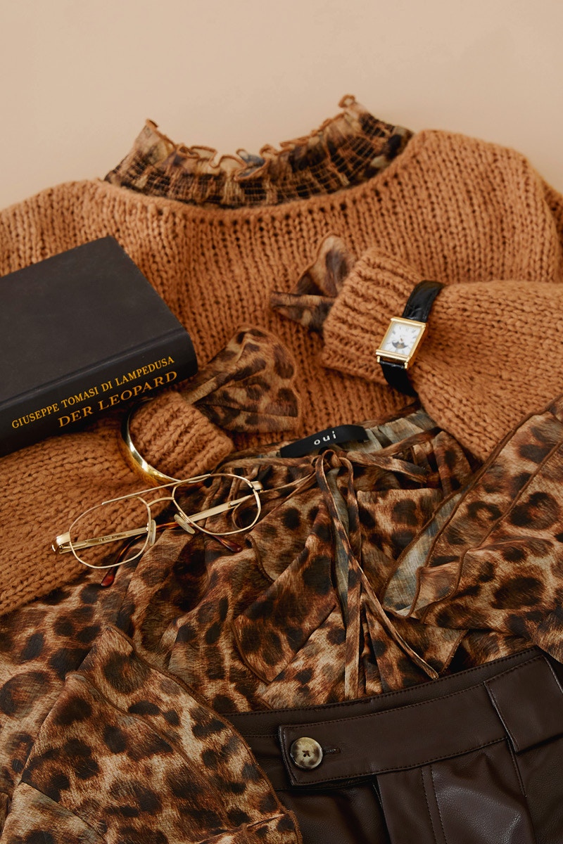 Close-up of a brown jumper under which a leo dress or leo blouse emerges, along with a wristwatch, a notebook in black and a pair of glasses.