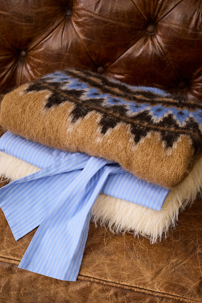 Folded clothes on a dark brown leather sofa. A Norwegian jumper in the colours brown, blue and dark brown on top, a blue blouse underneath and a plush white jumper at the bottom.