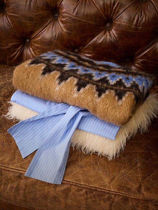 Gefaltete Kleidung auf einem dunkelbraunen Ledersofa. Oben auf ein Norweger Pullover in den Farben, braun, blau und dunkelbraun, darunter eine blaue Bluse und ganz unten ein plüschiger weisser Pullover.