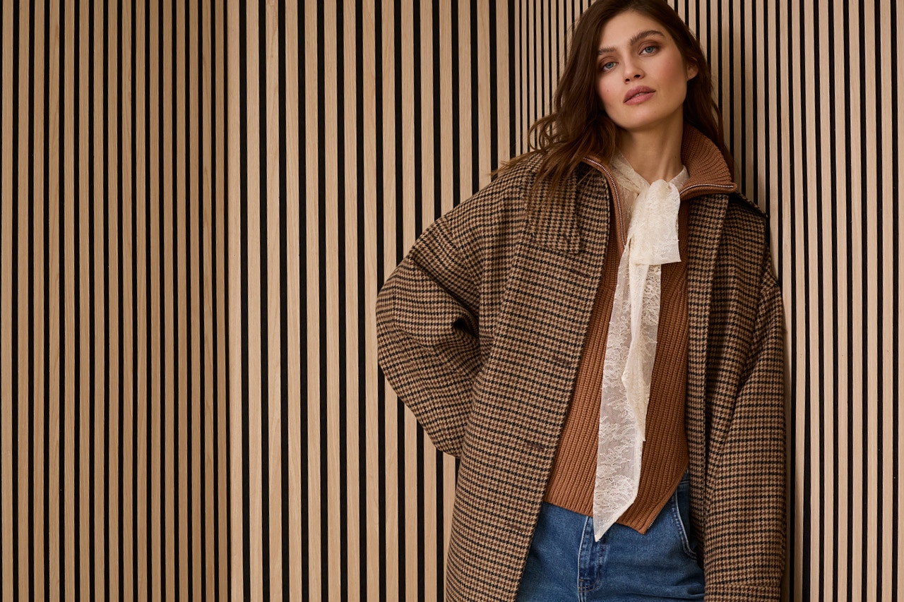 Woman wears a checked brown coat, a white lace blouse can be seen under a brown jumper and she is wearing jeans.
