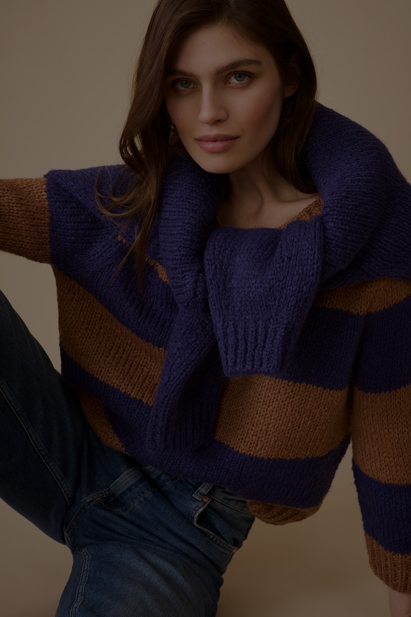 The woman is wearing a blue and brown striped jumper and has a blue jumper draped over her shoulders.