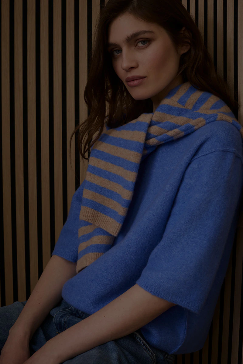 The woman is wearing a blue jumper and has a blue and beige striped jumper draped over her shoulders.