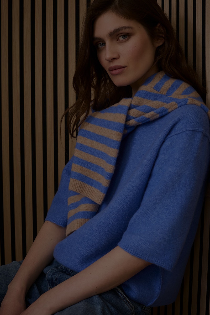 The woman is wearing a blue jumper and has a blue and beige striped jumper draped over her shoulders.