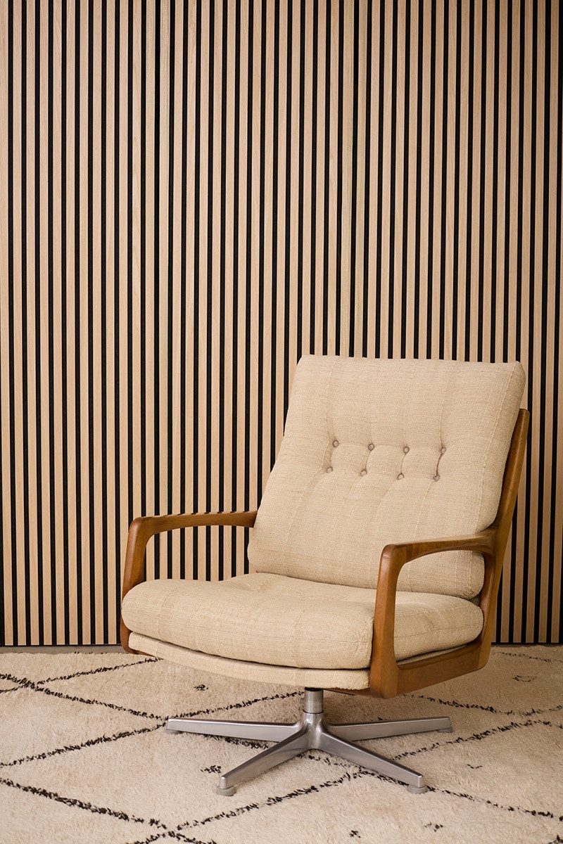 A white modern armchair on a white carpet with a large chequered pattern in front of a wooden panel.
