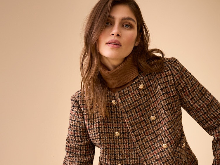 Woman wearing a brown checked jacket with gold buttons, with a brown turtleneck jumper underneath.