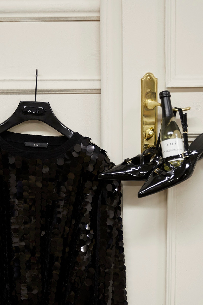 An einer Tür hängt ein paar Schuhe mit einer Prosecco Flasche an einem Bügel daneben hängt eine Jacke mit schwarzen Pailletten.