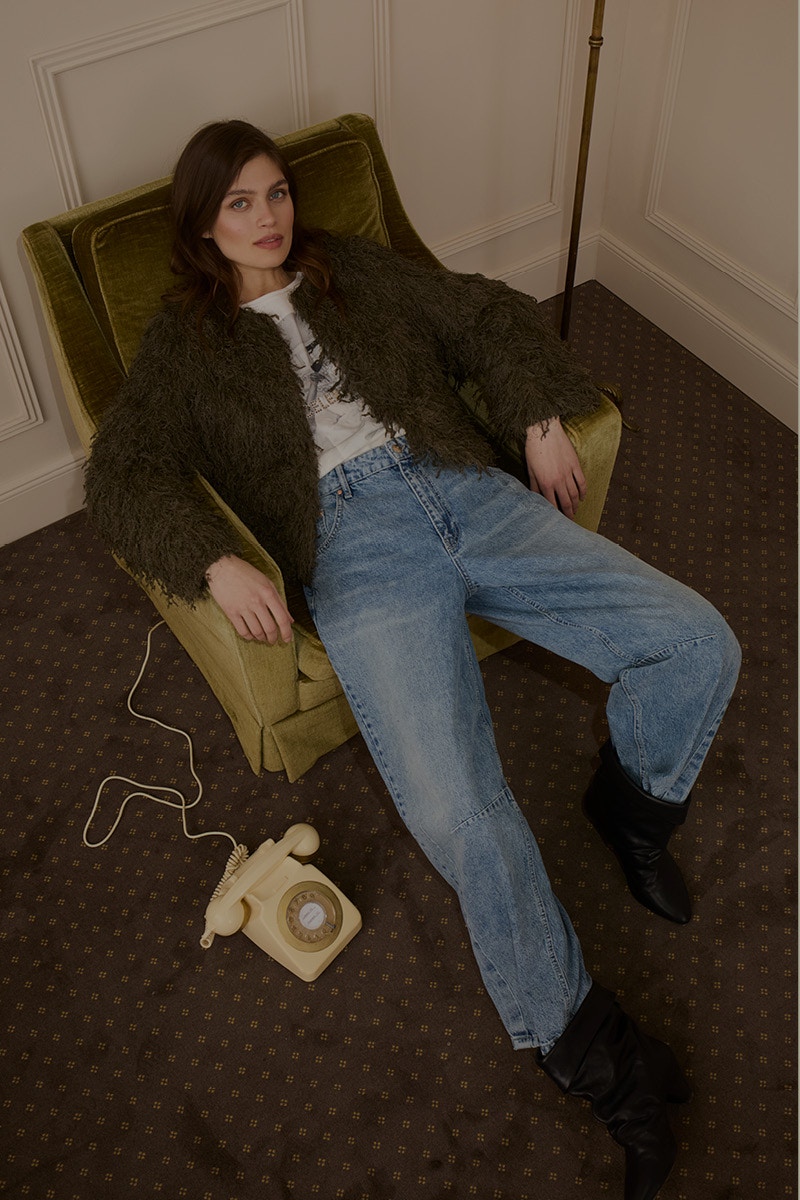 Frau sitzt in einem Sessel und trägt eine gruene Jacke mit einem gemusterten Shirt darunter und einer Jeanshose.