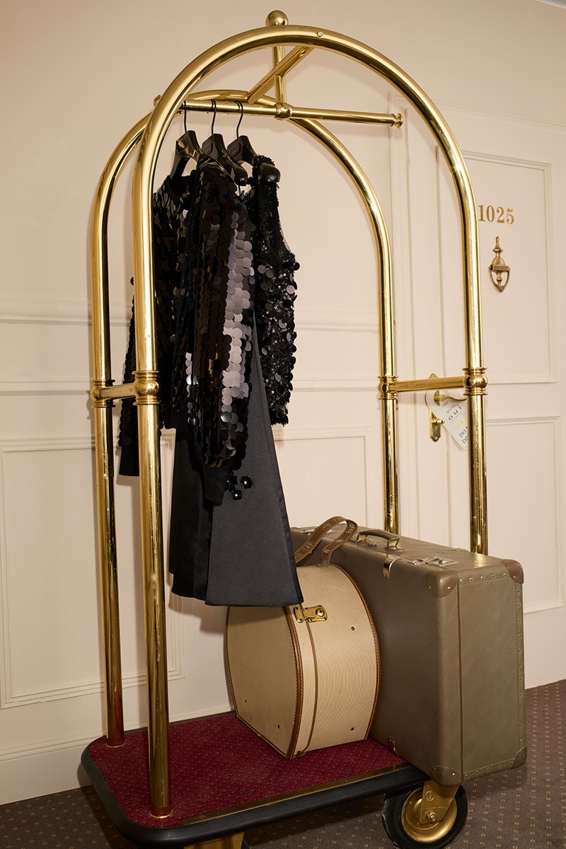 Gehängte Kleidung auf einem Hotelgestänge zum Transport von Koffern. Zu sehen sind mehrer Teile mit Pailletten eine graue Hose und 2 Koffer.
