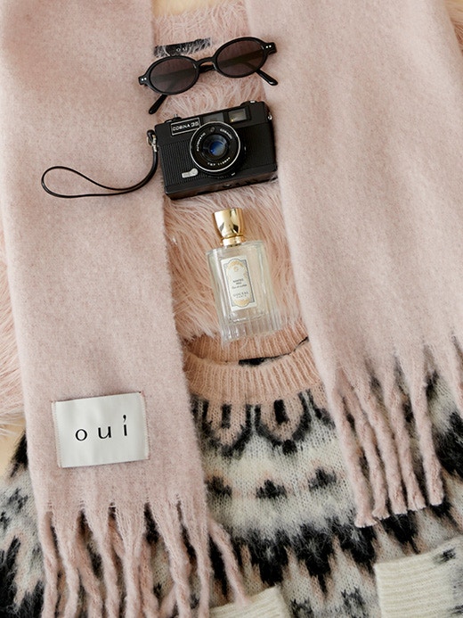 A jumper lies on the floor, it is patterned in black, white and pink. You can see the fringed ends of a pink scarf, a perfume, a camera and sunglasses.