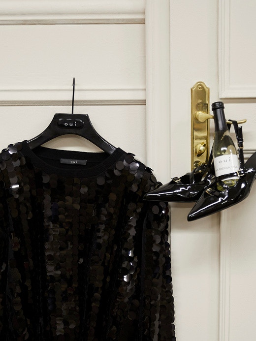 On a door hangs a pair of shoes with a Prosecco bottle on a hanger next to a jacket with black sequins.