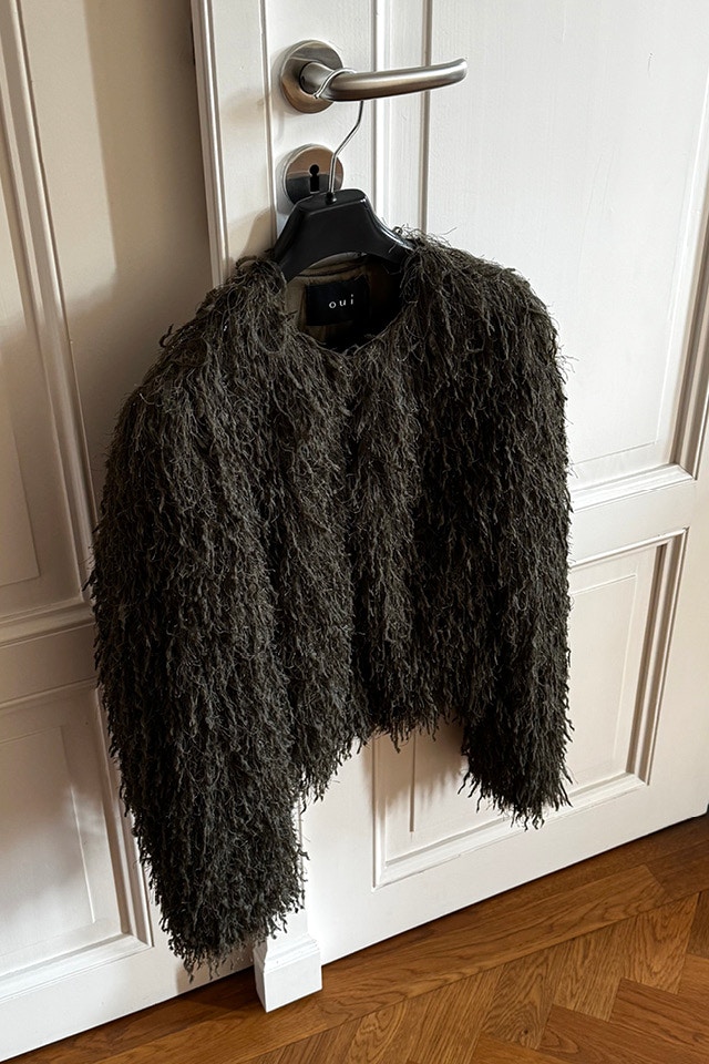 Eine dunkelgruene Fransenstrickjacke haengt an einem Kleiderbuegel an einer weissen Tuer.