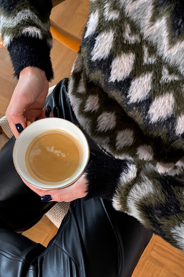 A close-up from slightly above, showing a seated woman wearing a fluffy jumper with a great pattern in green, black and white, and black leather-look trousers. The lady is holding a cup of tea.