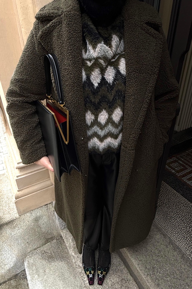 Man sieht einen Look an einer Frau aus einem khaki Mantel mit Strickpullover gemustert in schwarz, khaki und weiss dazu eine passende Hose in schwarz in Lederoptik.