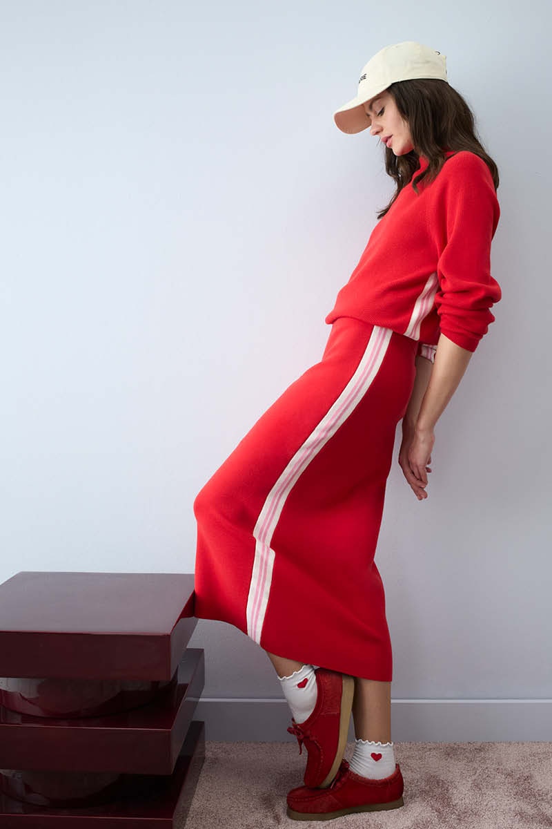 A woman is standing sideways, wearing a red jumper with a pink stripe down the side and a matching long red skirt, also with a contrasting pink stripe down the side.