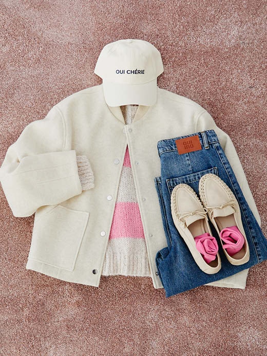 Ein Bild von Kleidung die auf einem plüschigen Teppich gelegt ist. Man sieht eine weiße Jacke im Collage-Stil darunter einen gestreiften Pullover in weiss mit rosa, eine gefaltete Jeanshose und weiße Loafer runden den Look ab.