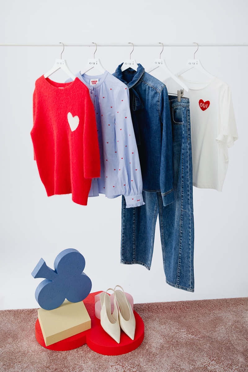A clothes rail with a red knitted jumper with a white heart on the chest hanging on it, behind it a striped light blue blouse, then a denim shirt, denim trousers and a white T-shirt with a red heart on the chest.