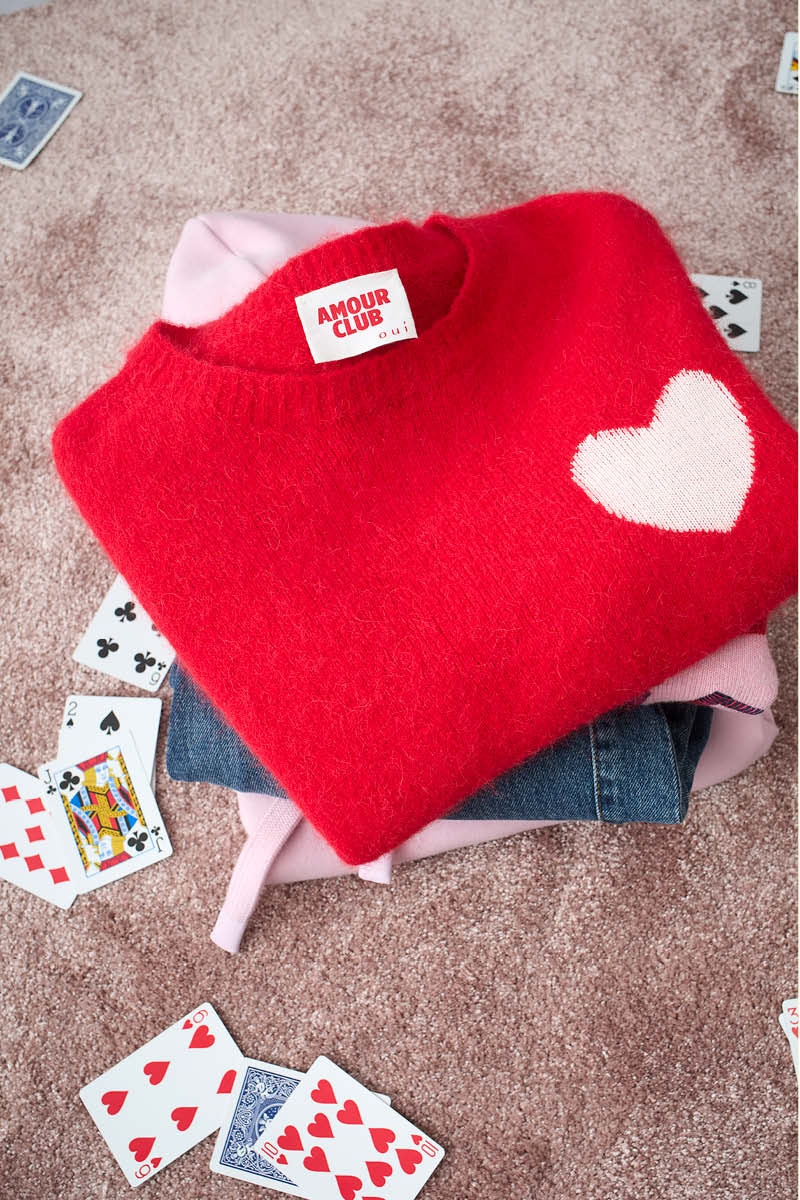 Ein Stapel Kleidung liegt auf einem plüschigen beigen Teppich. Man sieht eine Jeans, darüber einen rosafarbenen Pullover und oben auf einen roten Pullover mit einem weißen herz auf der rechten Brusthöhe, umgeben ist der Stapel von Spielkarten.